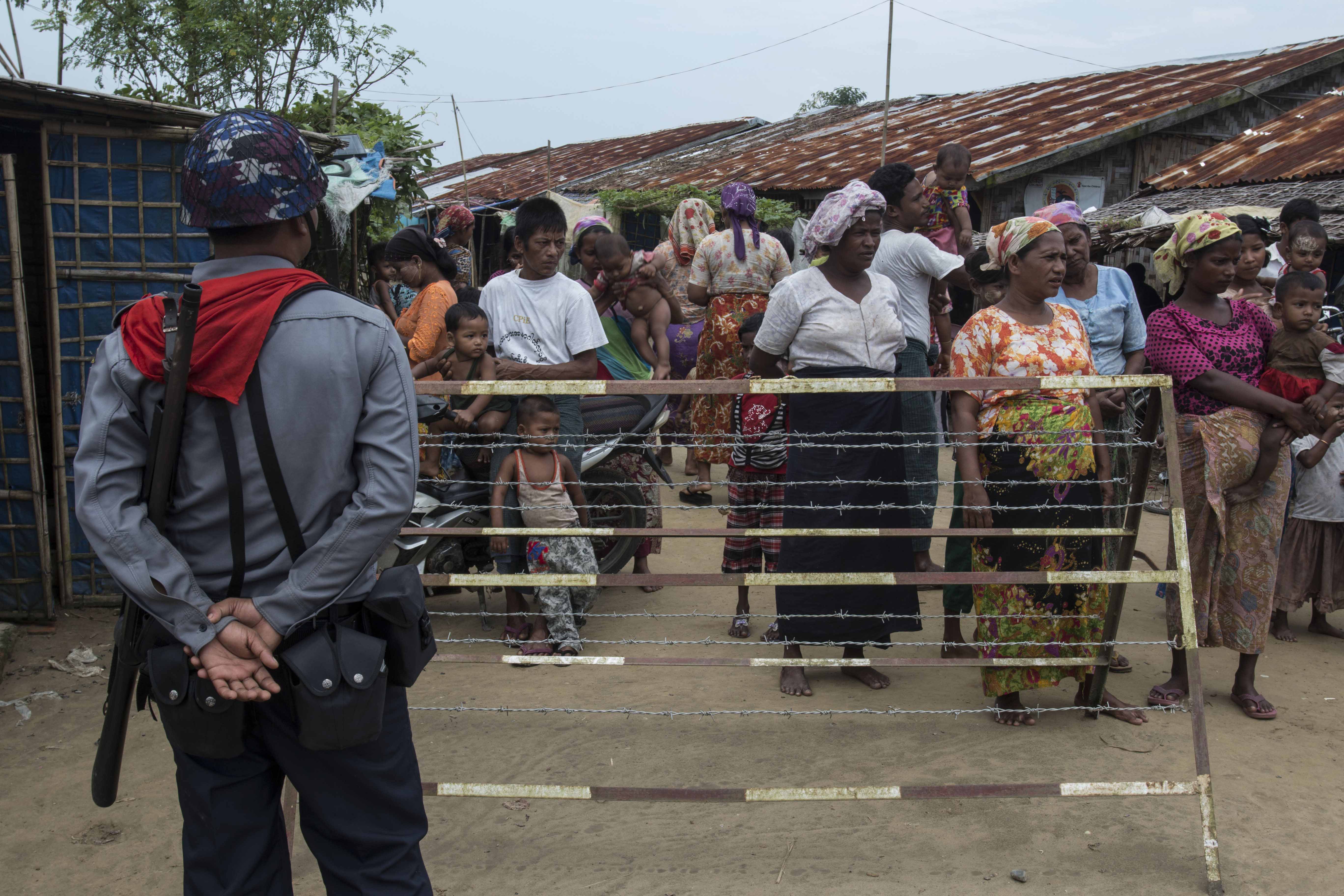 A crowd of people stand behind a fence while an armed officer watches them