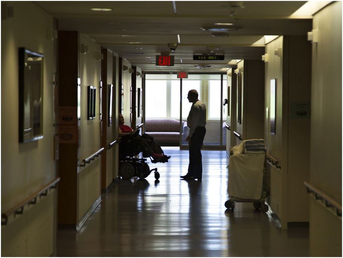 A woman in a wheelchair talks to a man who is standing in a nursing home.