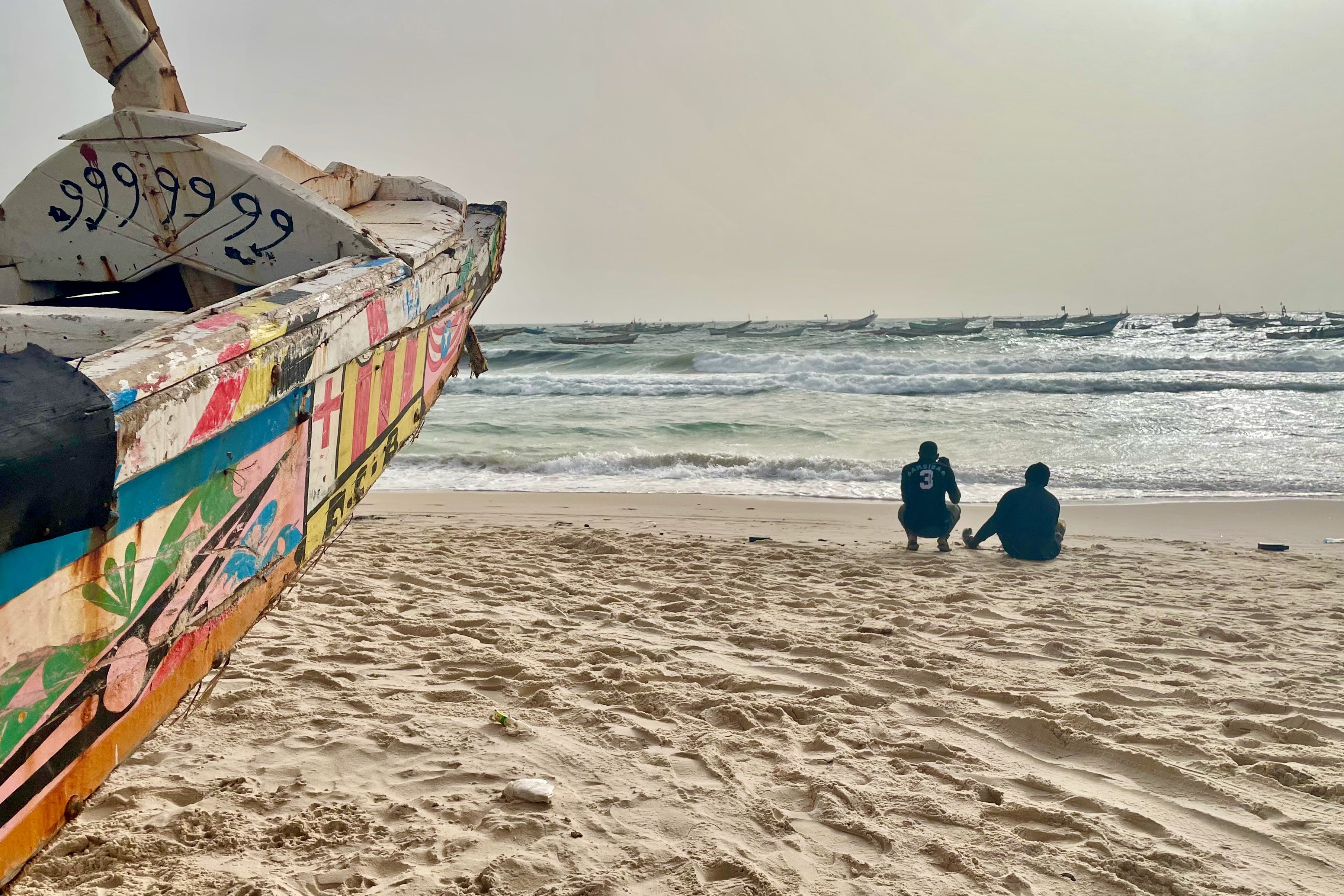 Two men sitting on a beach near wooden boats