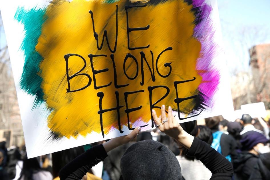 People demonstrate at a rally in Chinatown, New York City. Someone is pictured holding a painted sign reading "We Belong Here" with yellow, blue, pink, and black colors..