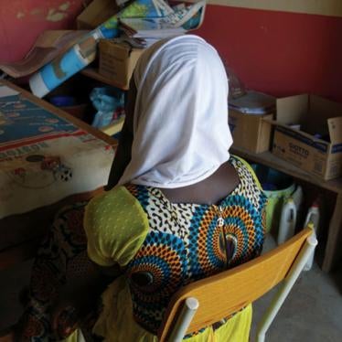 A girl sits in a chair, with her back facing the camera, in a colorful dress and a white headscarf.