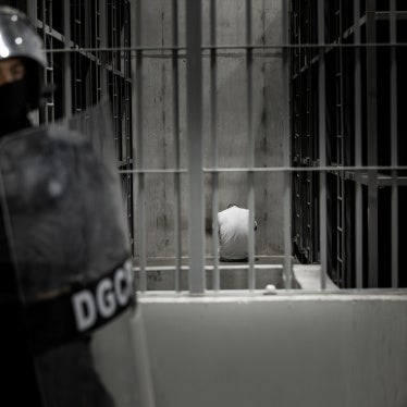 A person sits inside a cell while a guard with a riot shield stands outside
